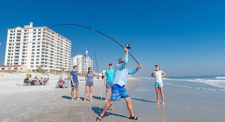 Man in blue shorts casts a fishing line in a wide arc on a sunny beach while others watch; high-rise buildings line the shoreline.
